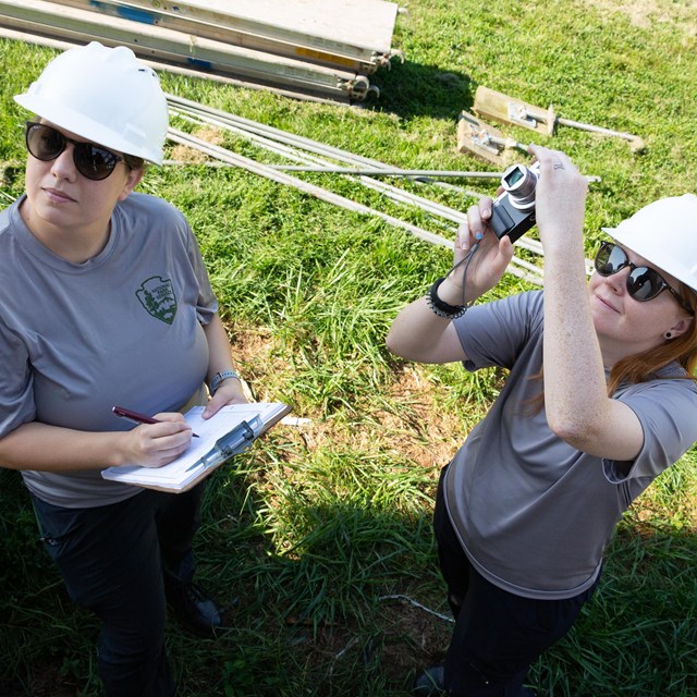 Two interns wearing NPS tshirts and hard hats look up while holding a clipboard and camera.