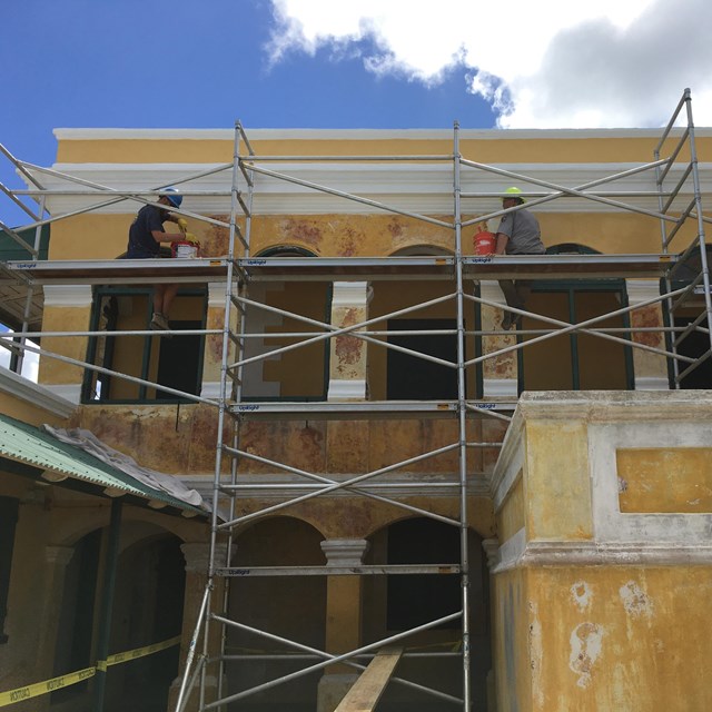 Crews work on tall scaffolding surrounding a bright yellow deteriorating wall.
