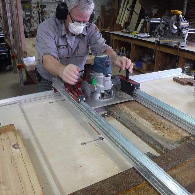 A person wearing a mask and ear protection uses a tool on a piece of wood.