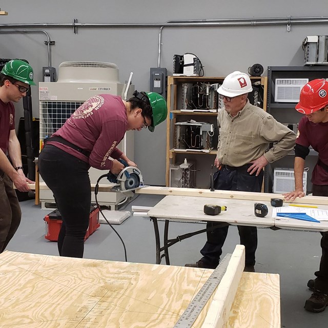 Four people at a workbench. One is using a saw while two hold down a board.