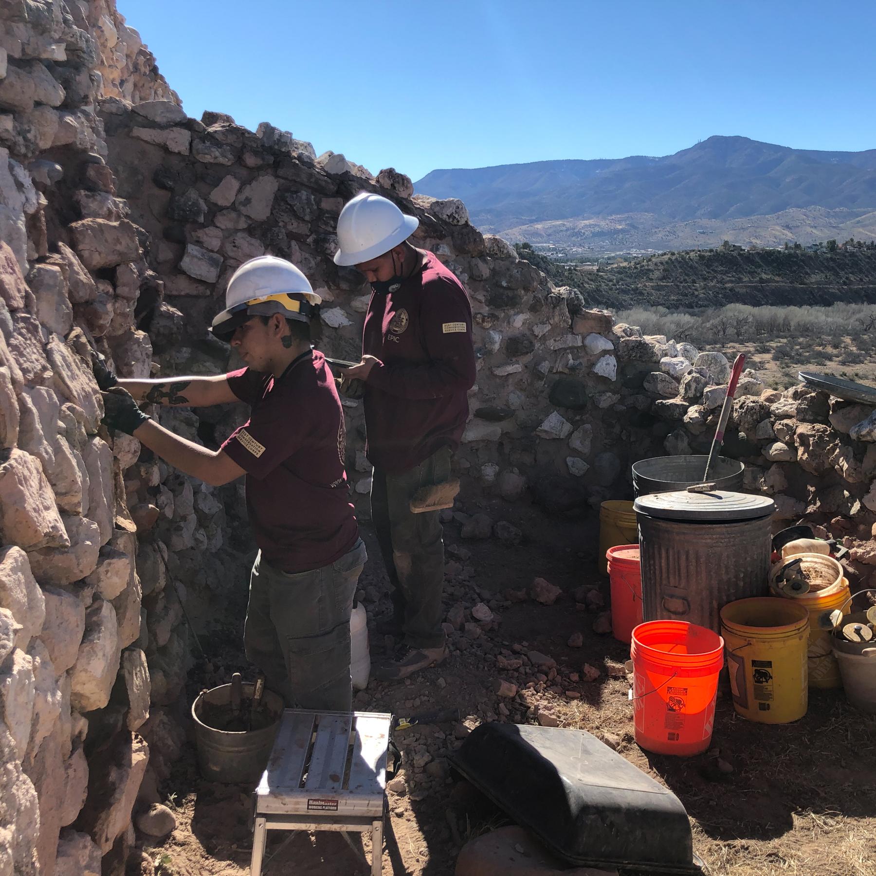 Two workers in hard hats and tool belts replace mortar between large stones in a prehistoric wall.