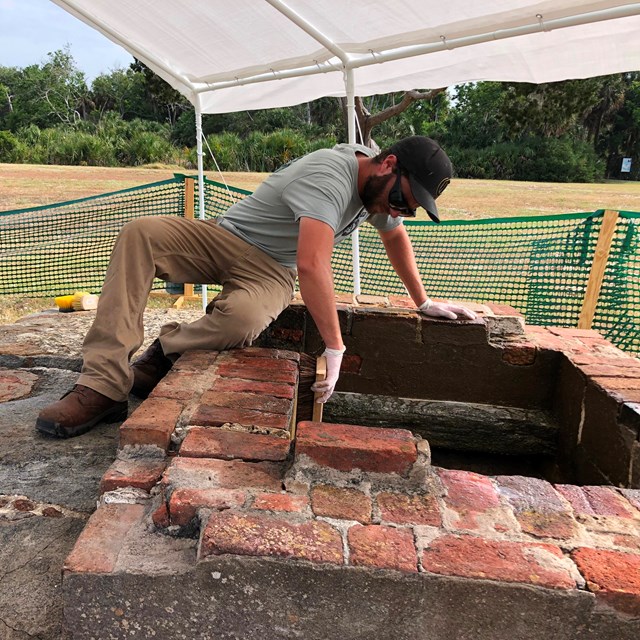 A worker under a sun shade works on a brick fireplace.