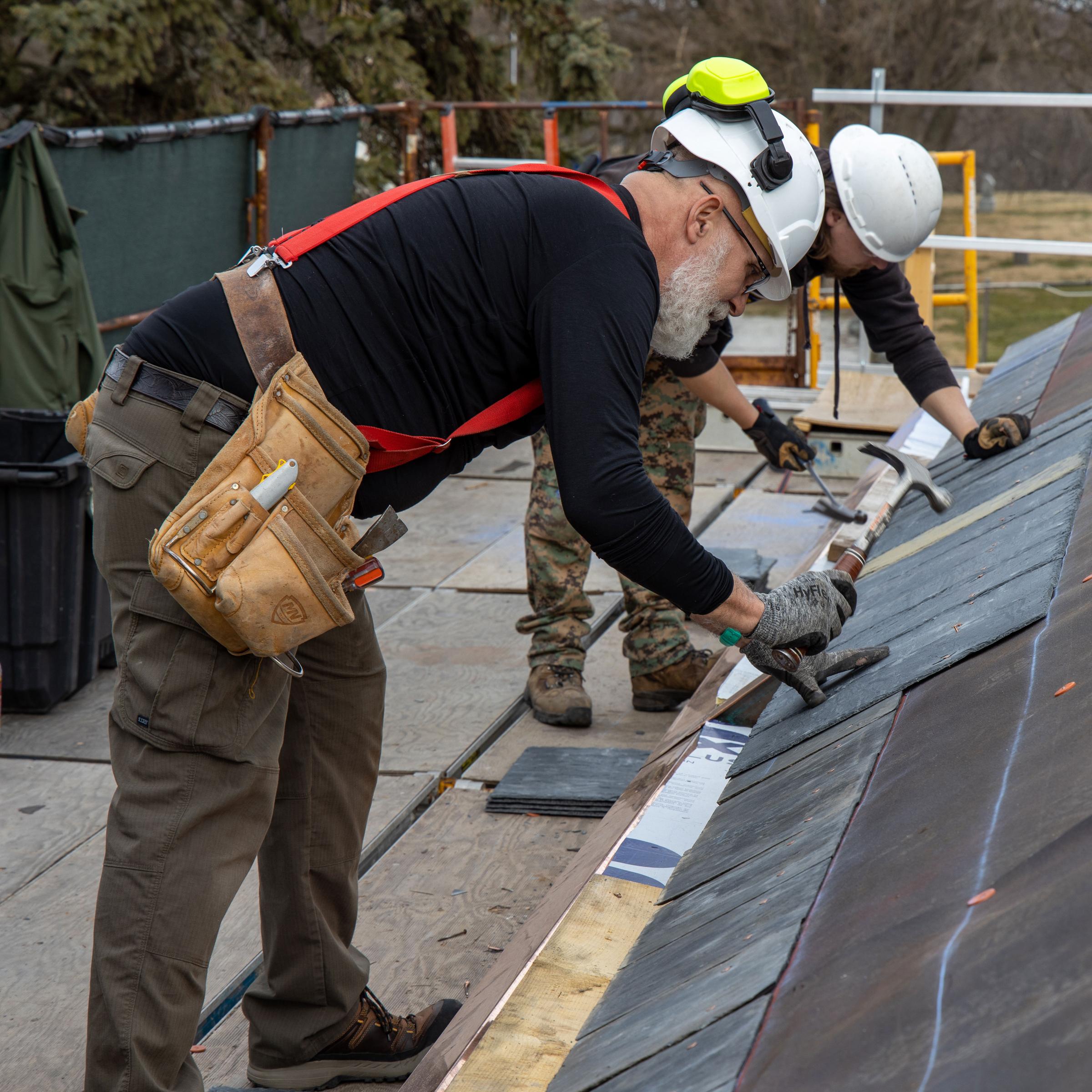Two workers in hard hats and tool belts nail slate tiles on a roof.