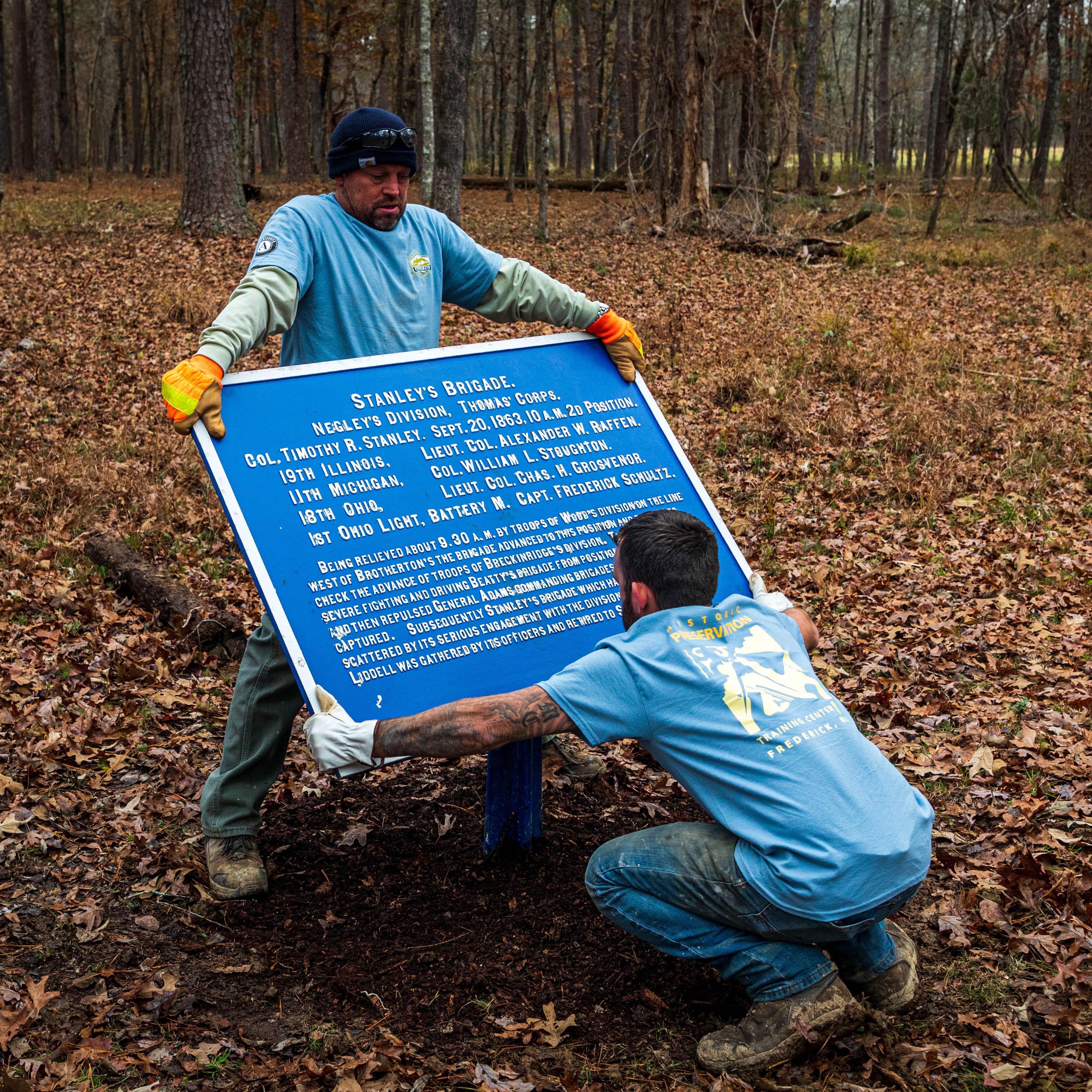 Two people lift a heavy metal panel describing a battle of the Civil War.