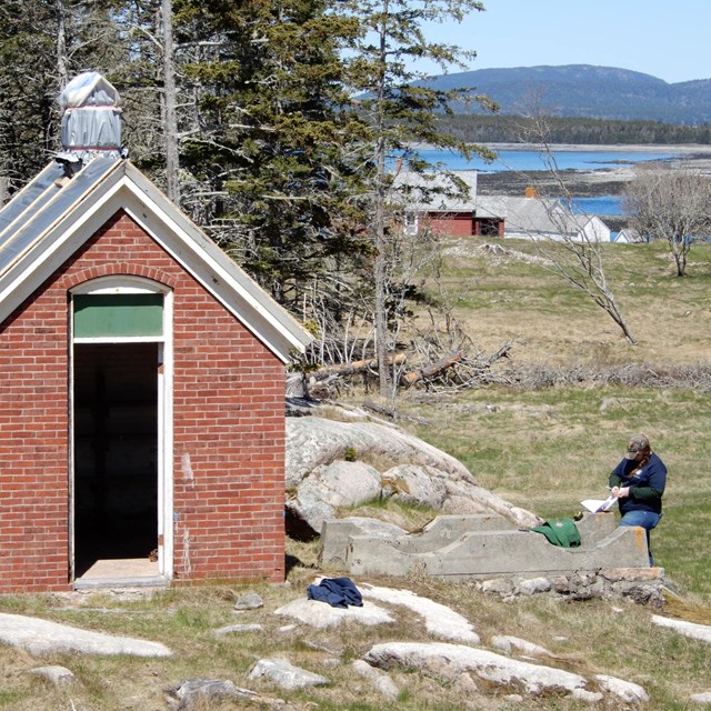 A person stands next to a one-room, brick building.