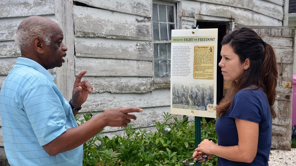 Two people stand in front of a run down building and discuss the history of the Underground Railroad