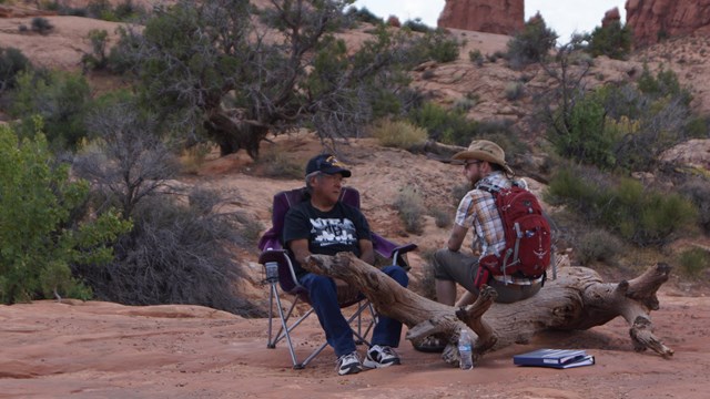 Two men sit, one in a chair and one on a log, in a desert area.