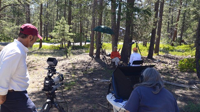 Two people film another interview a tribal member in a wooded area.