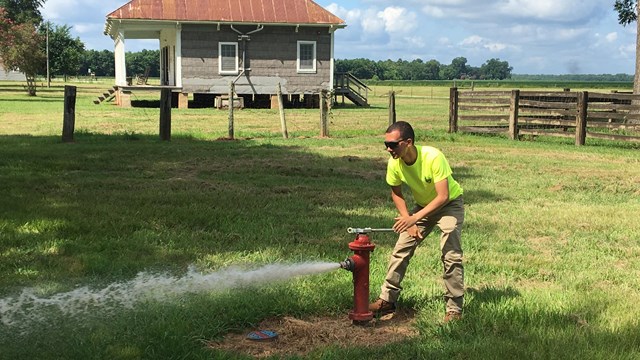A man flushes a fire hydrant in a field near an old structure.