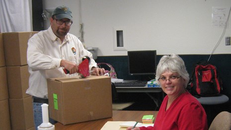Two people at a desk with box and documents in front of them.