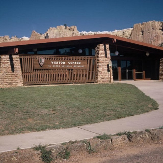 A wood and brick building with peaked roof in front of a cliff face.