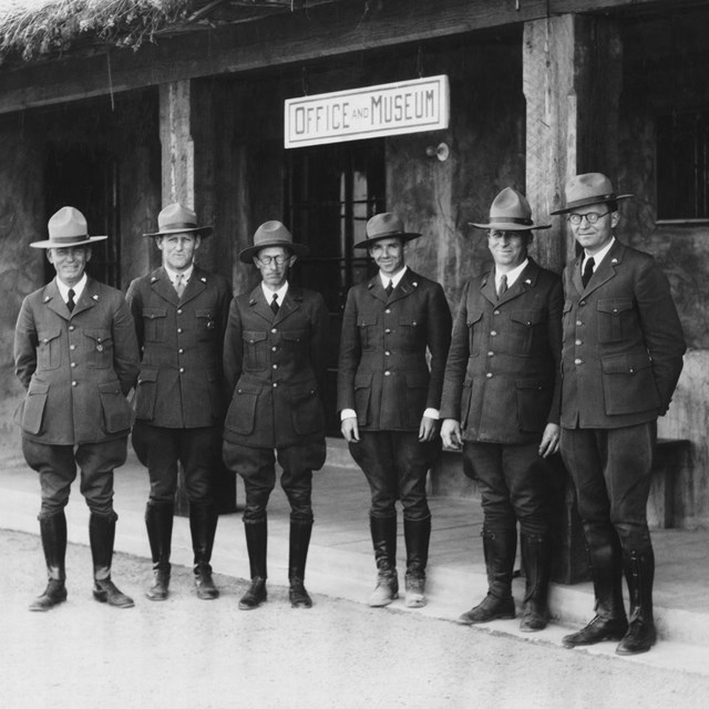A group of 6 men stand in early NPS uniforms in front of an adobe building with thatch roof.