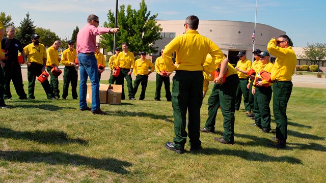 A man in a red shirt points while men in fire gear look on.