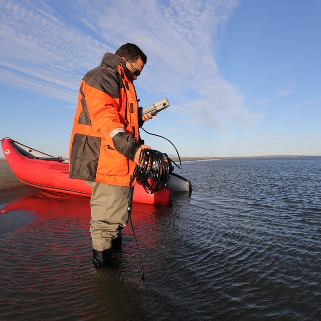 A scientist in an orange float coat next to an inflatable raft takes water quality measures.