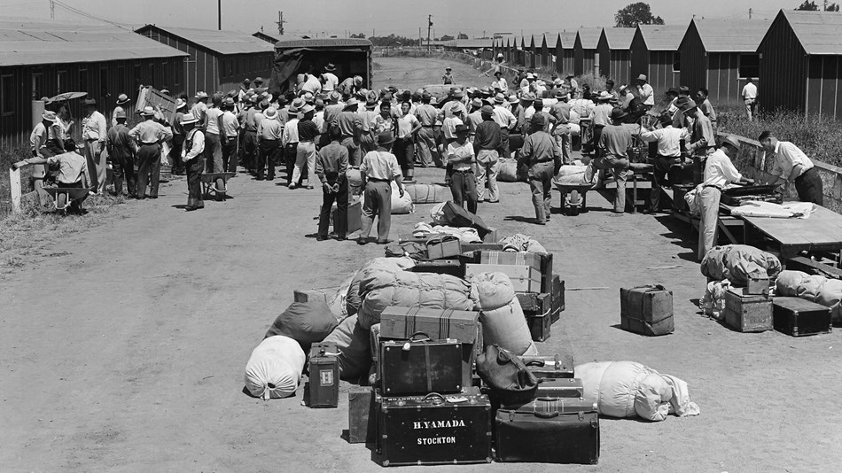 historic photo of a large group of men loading suitcases onto a truck surrounded by barracks.