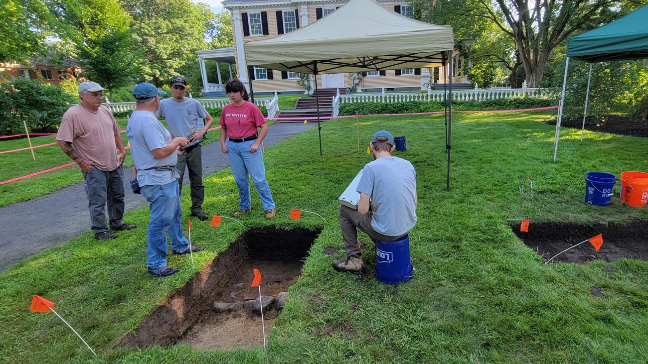 Five archeologists chat about the nearby test unit in front of a large yellow house. 