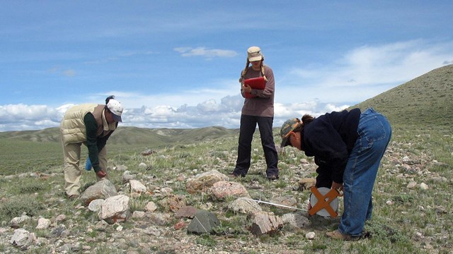 three people doing a measurement over a rock pile, grassy hills around, blue skies