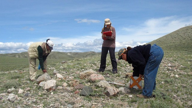 Three people bend over, measuring a rock grave, grassy hills, blue skies