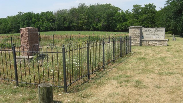 grass in foreground, fenced in cemetery, rock site identification sign: Santa Fe Trail