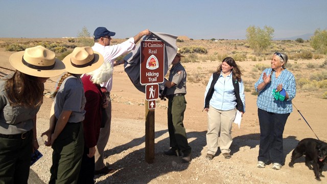 people outside standing on a road unveiling a trail sign, one black dog on leash