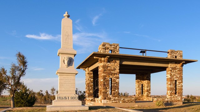 Tall, carved stone monument next to a covered shelter with columns or rock on top of a hill.