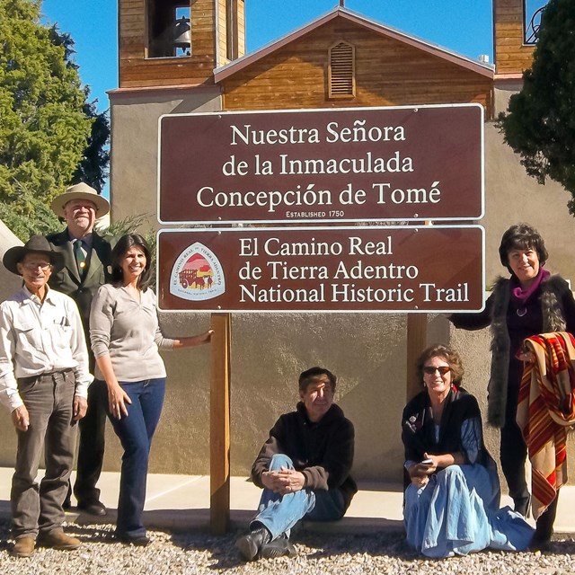 People standing and sitting next to a brown sign with white text in English and Spanish