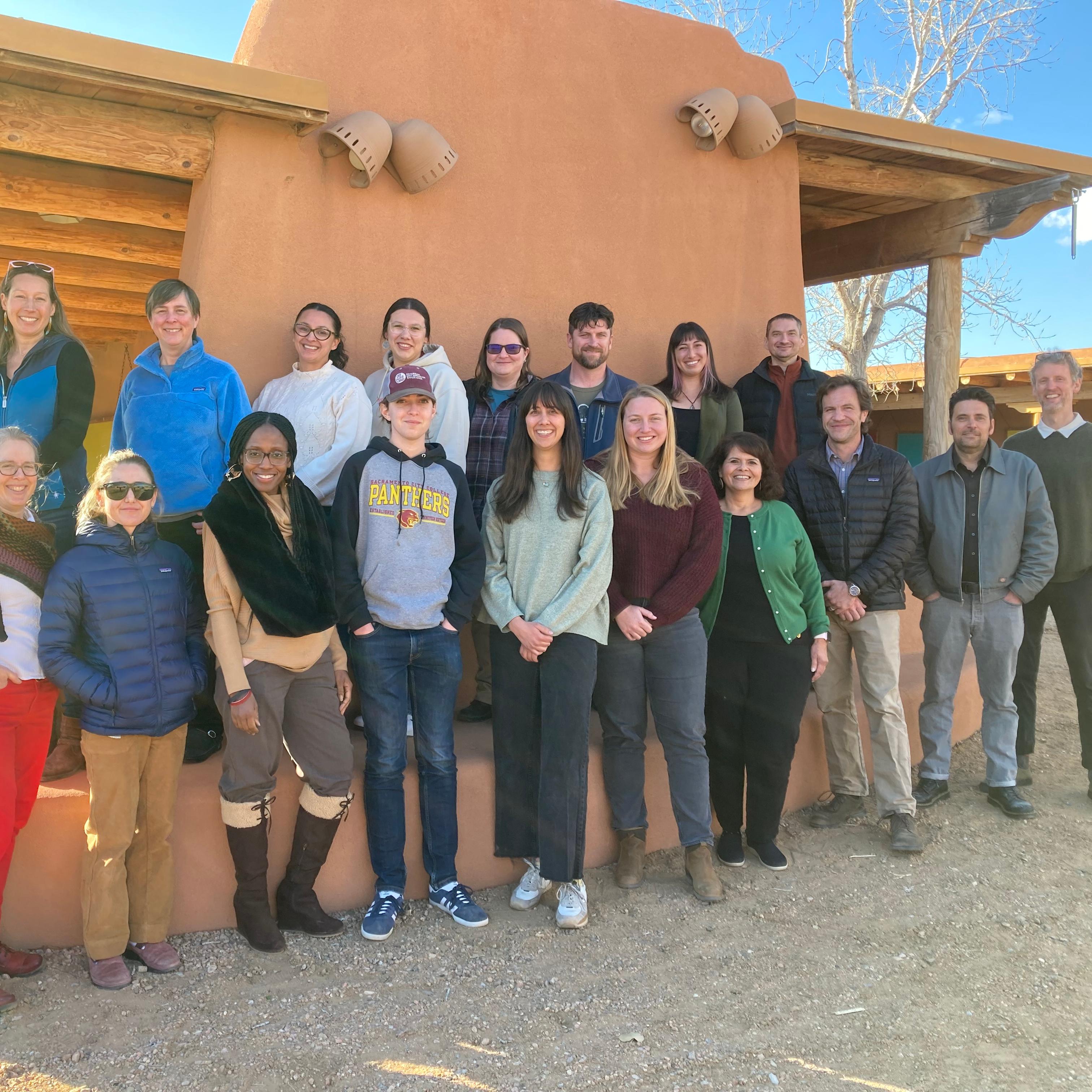 A group of about 20 people stand for a photo in a desert setting next to a wall.