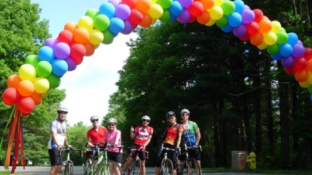 A group of bicyclists under a banner of rainbow balloons.