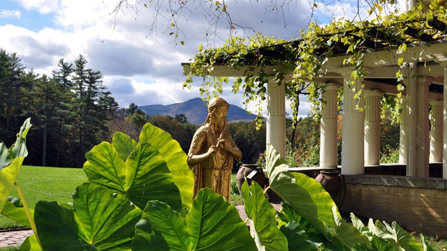 A gold statue behind leafy foliage, with a colonnade and mountain in the distance