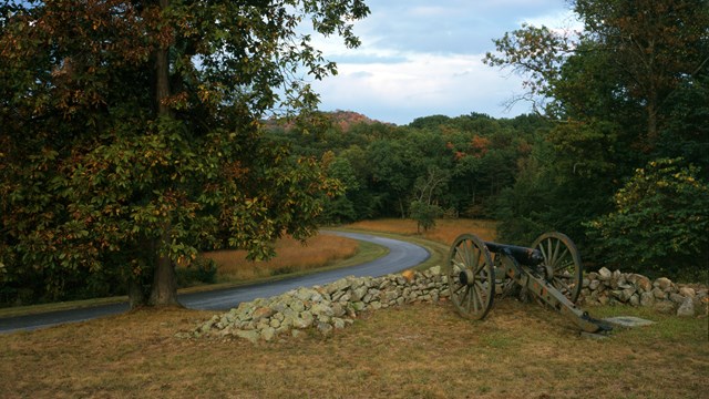 A cannon stands behind a low stone wall in a rural landscape of wooded areas and open grass