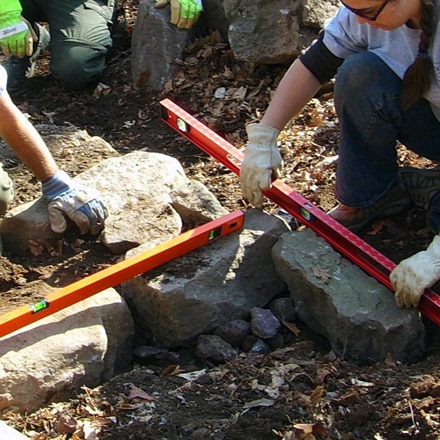 A group of three people use levels to place boulders along a pathway.