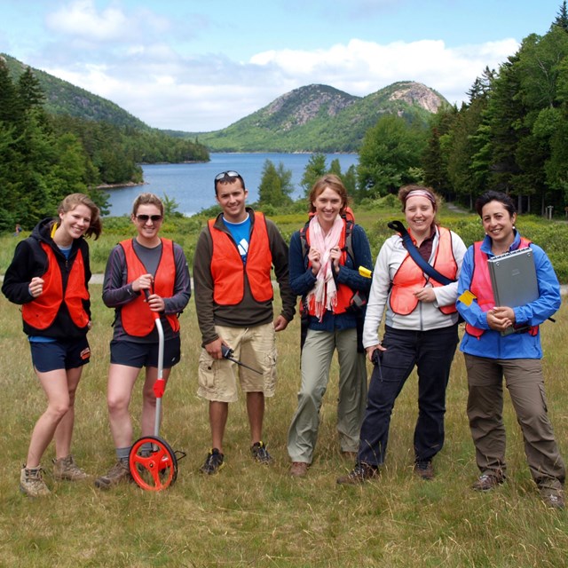 A group of six young people with work gear pose in front of a view of a lake and two round hills.