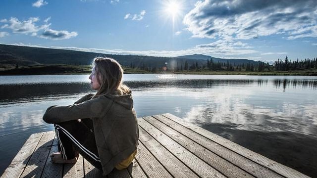 A woman sits on a dock near reflective water with bright sky above.