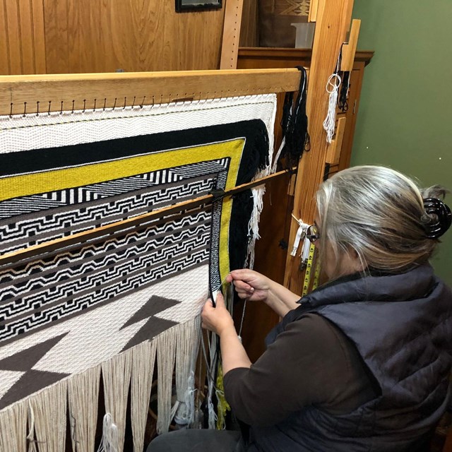 A woman sits at a side shot loom weaving. Photo credit: Lani Hotch/Chilkat Indian Village