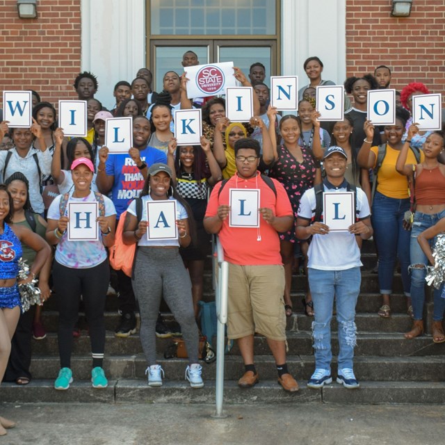 Men and women stand in front of a brick building holding signs that read Wilkinson Hall