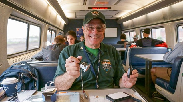Senior male volunteer sits at a table on a train, while holding an NPS passport stamp. 