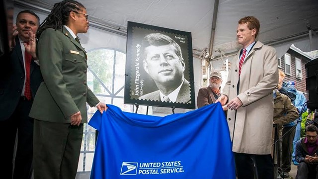 Woman and man are unveiling a large photo of the new John F. Kennedy postage stamp.