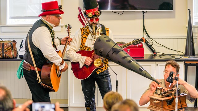 Men in odd clothing record a song onto a wax cylinder