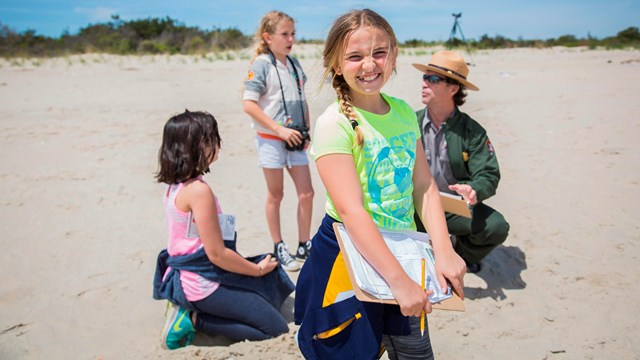 Three young girls are on the beach with a park ranger. One girl is holding a clip board and smiling.