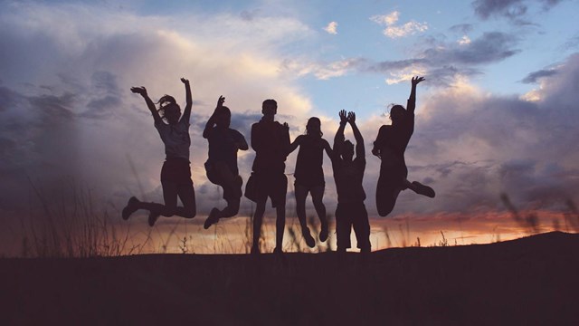 Silhouette of six people jumping with cloudy sky behind illuminated by the setting sun. 