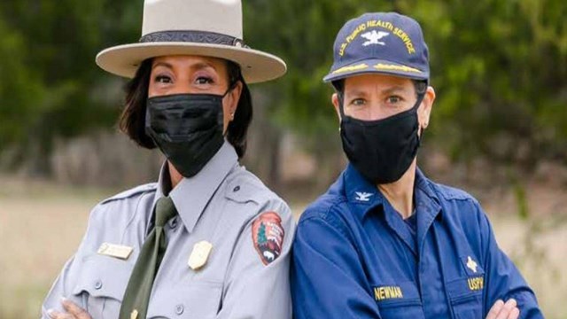 one person poses in a grey national park uniform next to a person in a blue USPHS uniform