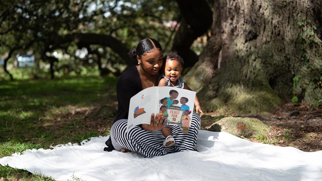 mother sits under a tree while reading to her baby in a park