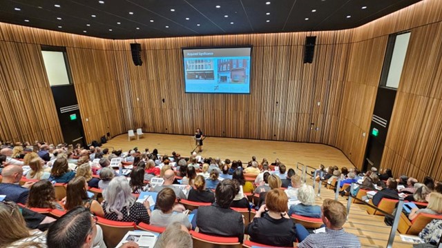 a woman stands at a podium in front of a projection screen in an auditorium filled with an audience