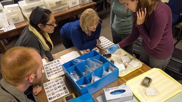 5 people gathered around a table scattered with papers & objects & a box of artifacts in the center