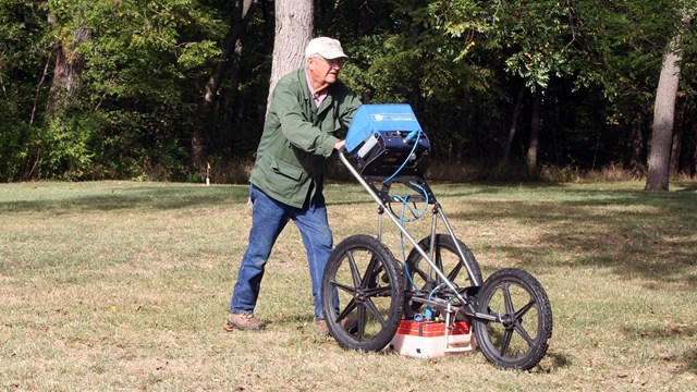 Volunteer operating ground-penetrating radar equipment in a clearing with trees in the background