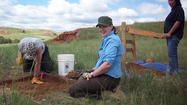 3 people digging a square test unit in a grassy field with large clouds in a blue sky above