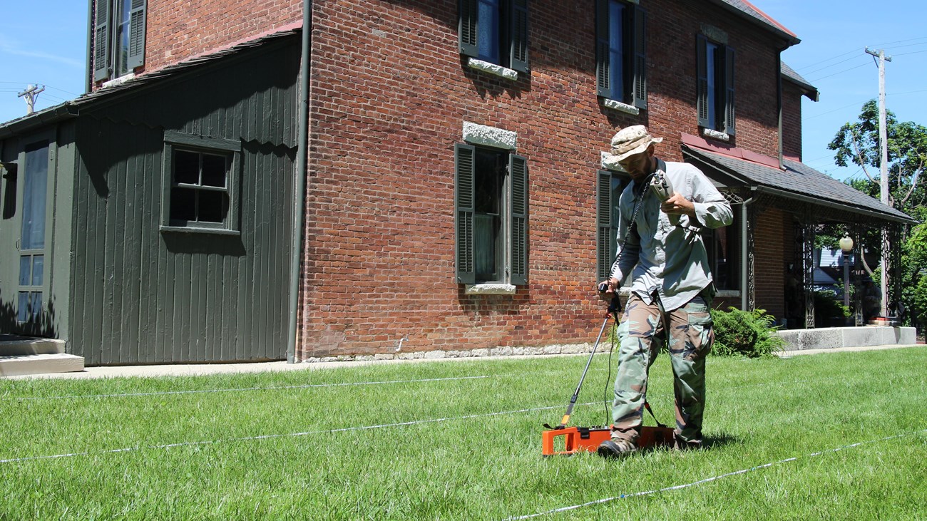 Archeologist holding a remote sensing instrument and walks across a lawn in front of a brick house