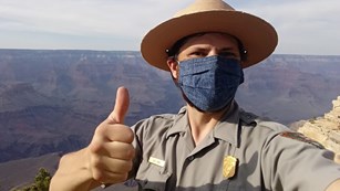 A man giving a thumbs up in ranger uniform and mask, in front of the Grand Canyon