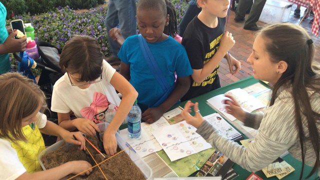 Kids dig in a small sandbox while NPS employee shows them a fake pottery artifact.