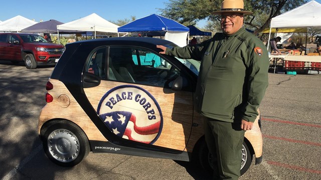 A man in a green uniform and hat stands in front of a tiny car.